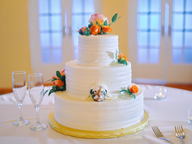 Three tier white cake with orange flowers and a dog peeking out of the first tier. 