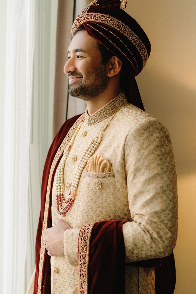 The groom smiles by a window while wearing traditional Indian wedding attire.