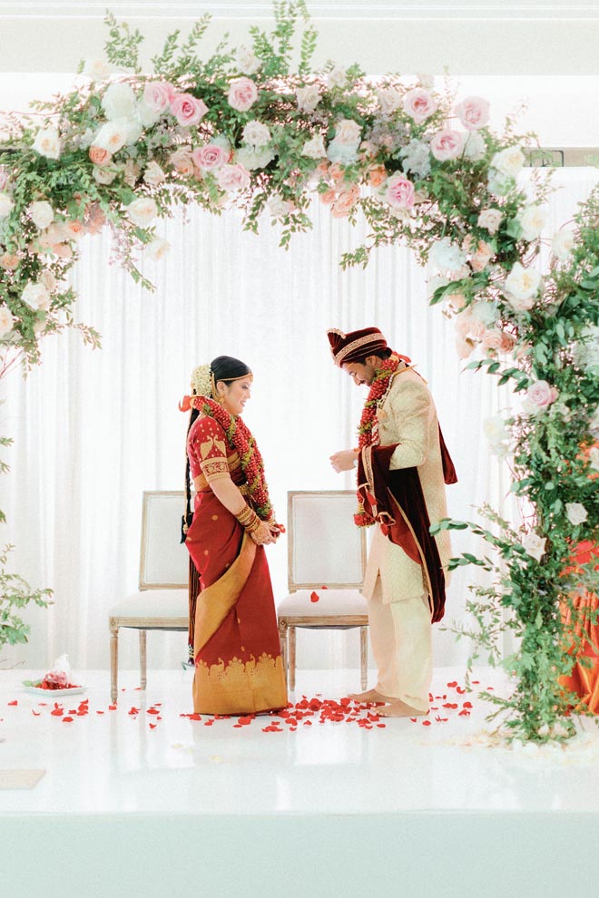 The bride and groom stand under a floral arch at their Hindu ceremony in Houston.