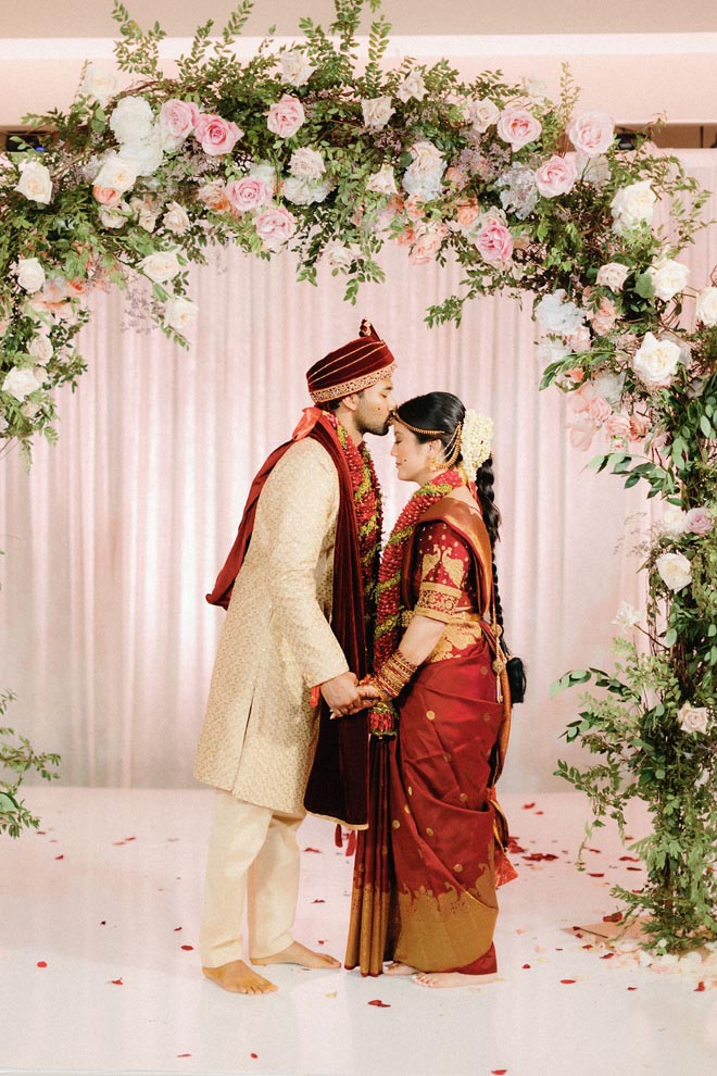 The groom kisses his bride on her forehead while standing under a floral arch at their traditional Hindu ceremony.