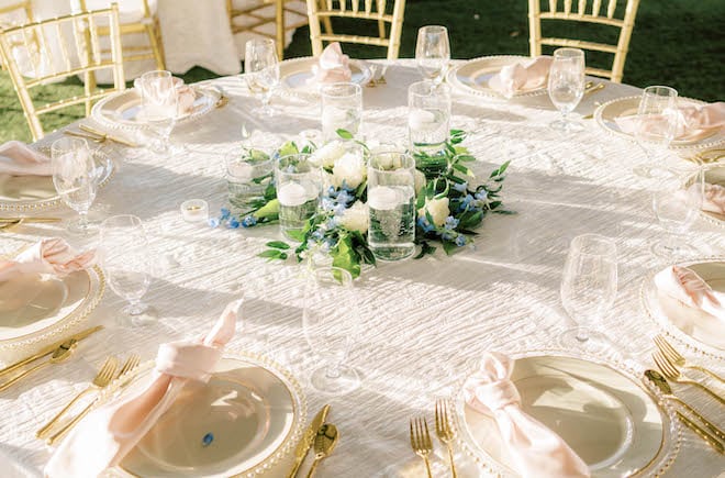 Blush napkins, gold silverware and plates, white roses, blue flowers and candles decorate the reception tables. 