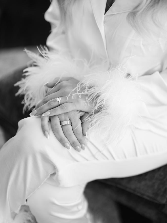 The bride wearing satin pajamas with feathered sleeves with her hands on her leg, showing her rings. 