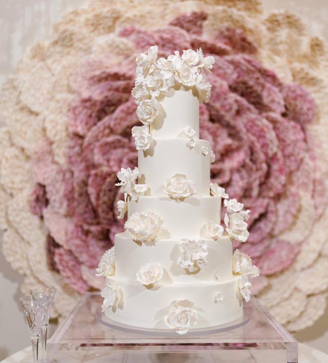 A five tier white cake with white floral garnishes against a neutral hued floral background. 