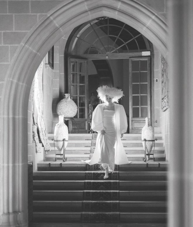 The bride walking down stairs in her wedding gown and a white feather hat. 