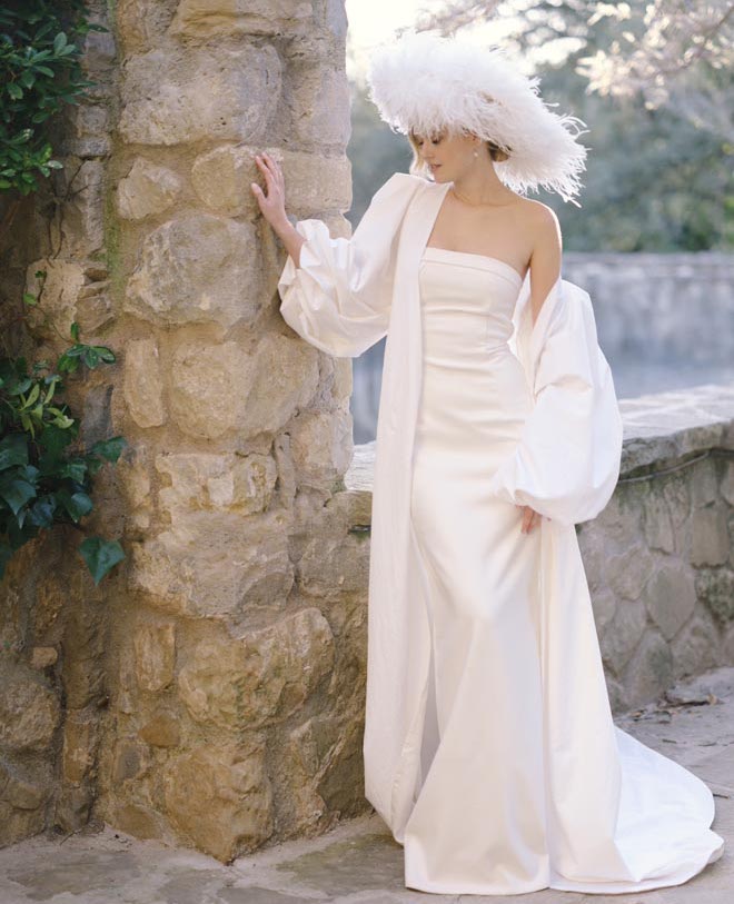 The bride leaning against a brick wall in her wedding gown and feathered white hat. 