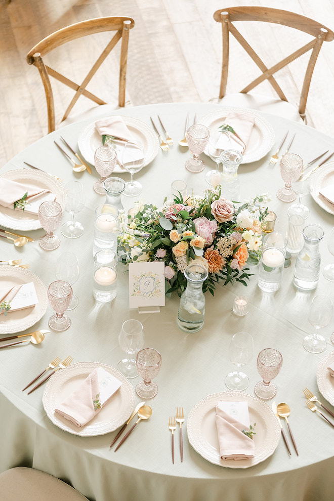 A round table with a light green tablecloth, pink chargers and gold flatware with a floral centerpiece. 
