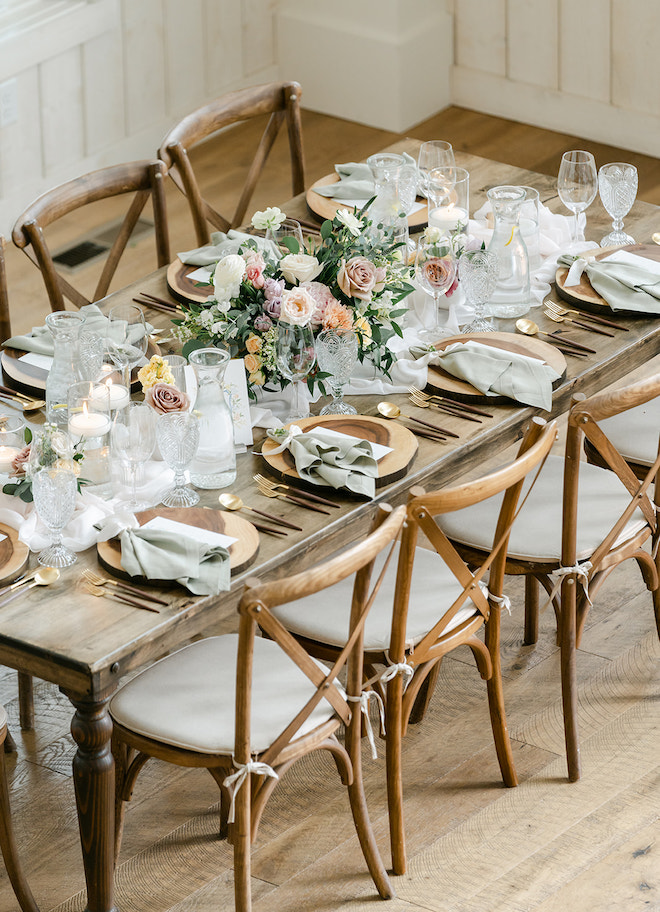 A wooden table set with gold flatware, green linens and florals. 