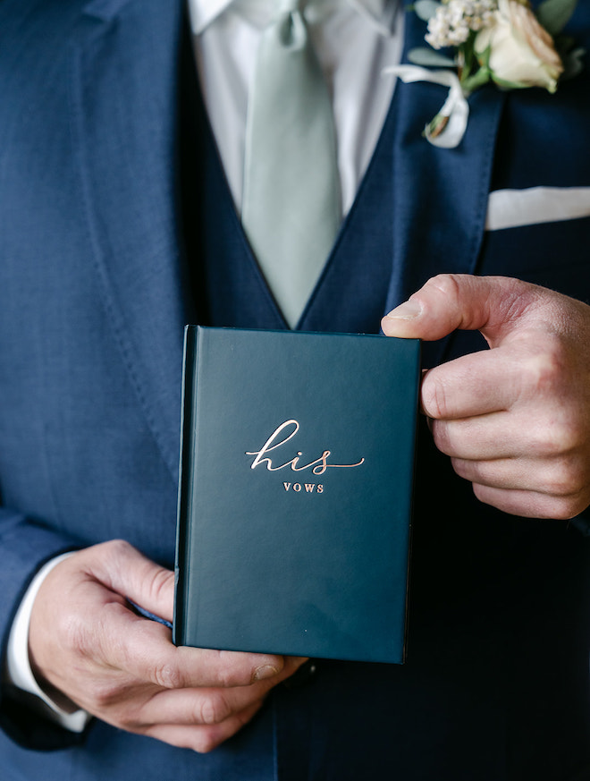 The groom holding a navy booklet with "his vows" written in gold on it. 
