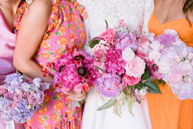 The bride and bridesmaids in pink and orange dresses holding their pink and purple floral bouquets. 