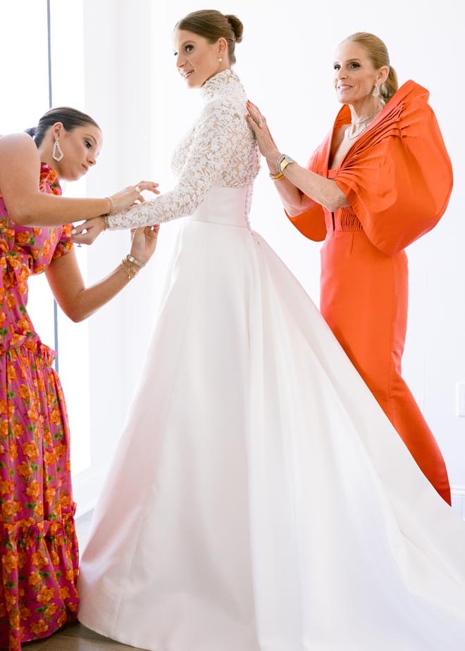 A bridesmaid and the mother of the bride wearing bright pink and orange dresses helping the bride get ready. 