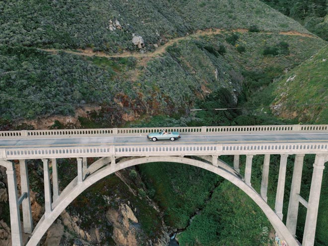 An ariel photo of a vintage blue Mercedes driving on a bridge over the California coastal mountains. 