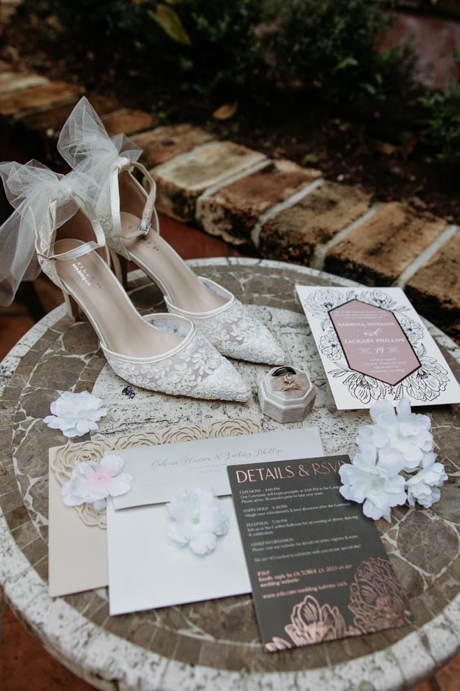 The brides shoes, wedding band and wedding stationery lay on top of a table outside at The Bell Tower on 34th.