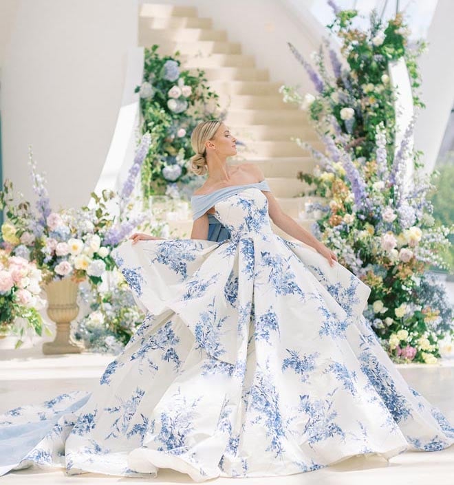 A bride wearing a blue and white printed wedding gown in front of large floral arrangements lining the stairs. 