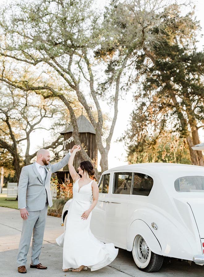 The groom spins the bride around a vintage white car. 