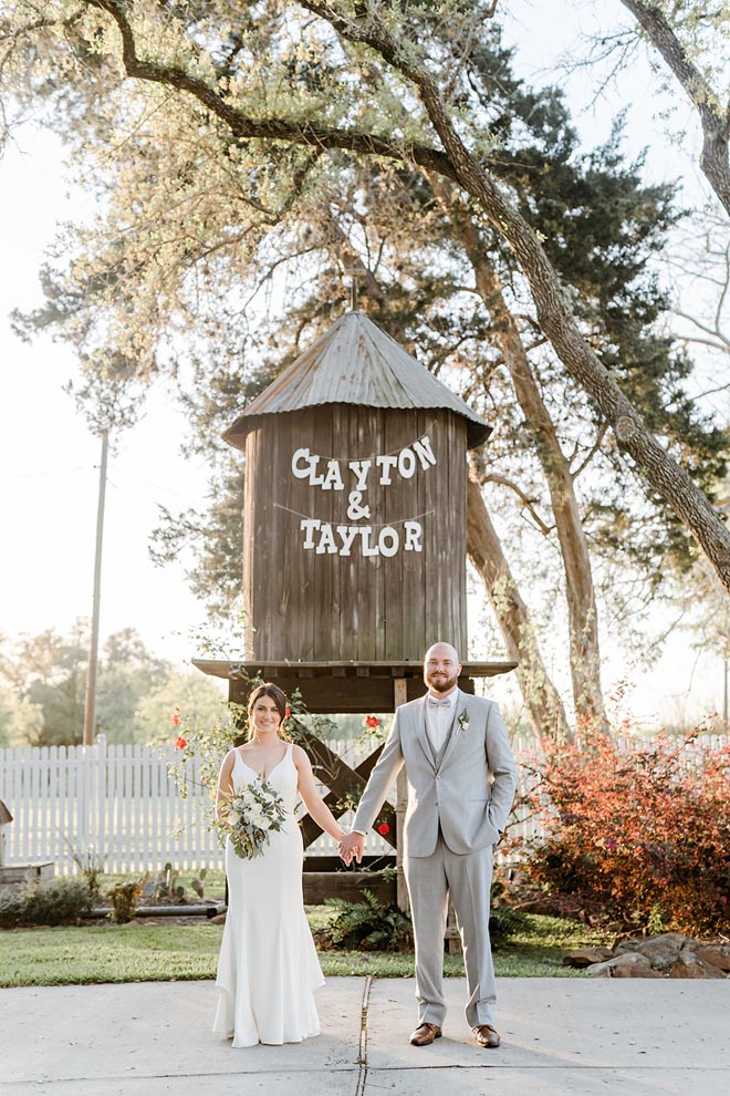 The bride and groom hold hands in front of a wooden tower with their names "Clayton and Taylor" strung across in decorative lettering. 