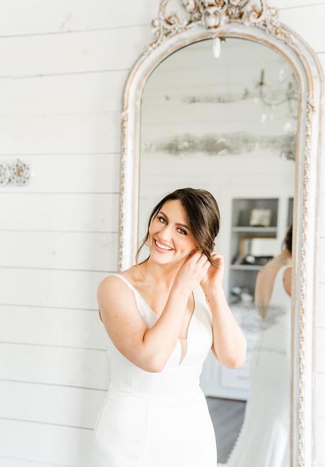 The bride smiles as she puts on her wedding day earrings before her French blue outdoor wedding ceremony.