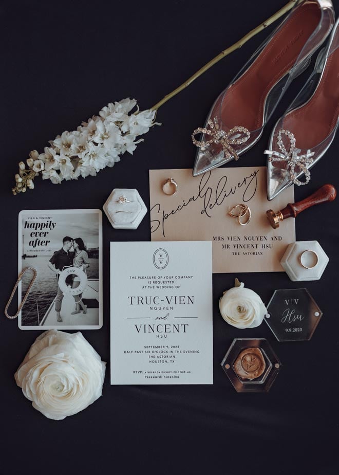 The wedding stationery, brides shoes, jewelry and wedding rings lay on a black backdrop.