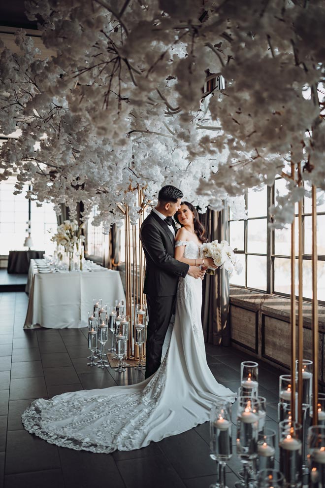 The bride and groom stand under a white floral instillation at their wedding venue, The Astorian.