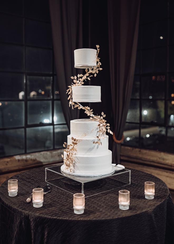 A five-tier white wedding cake detailed in gold ivy sits on an acrylic cake holder.