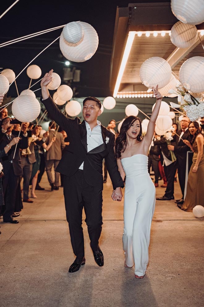The bride and groom hold hands as they exit their wedding reception under paper lanterns.