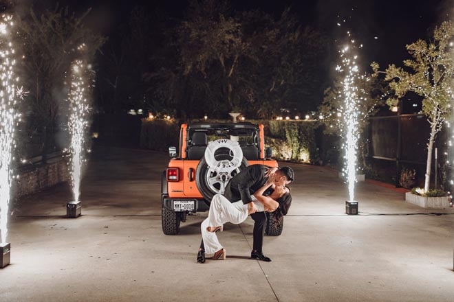The bride and groom share a kiss in front of an orange Jeep and sparklers.