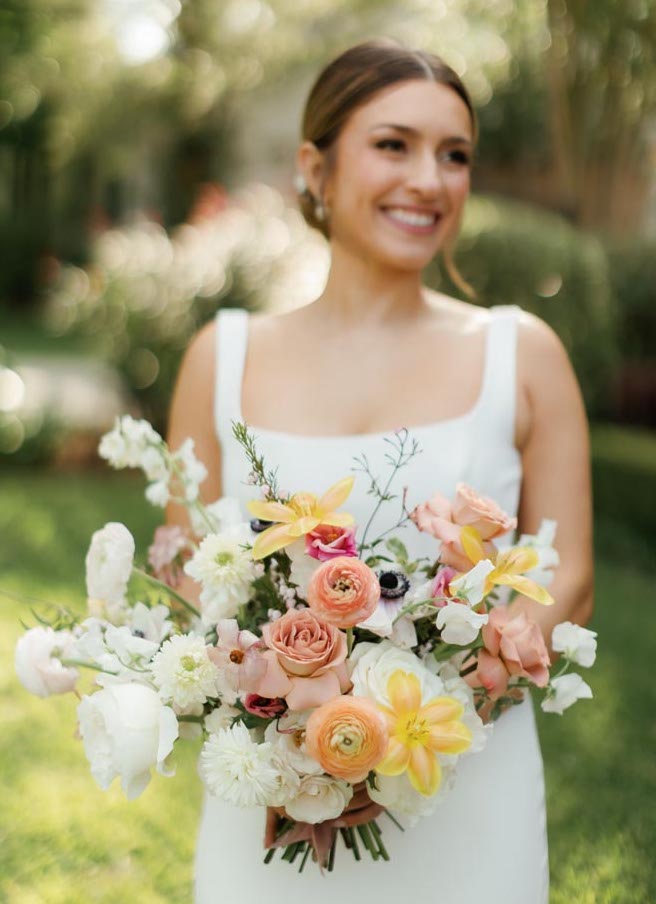 The bride smiles while holding an orange, yellow, white and peach floral wedding bouquet.