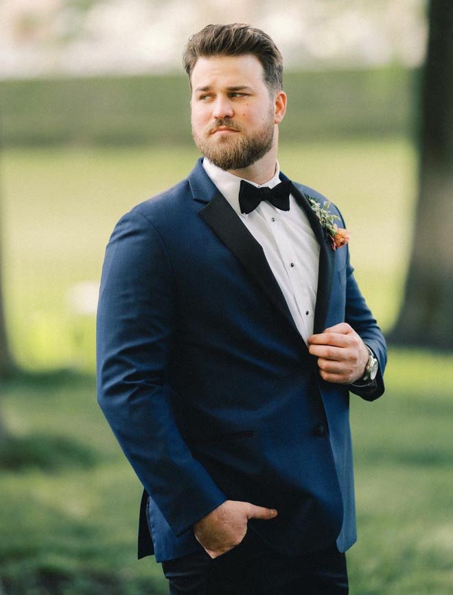 The groom stands outside the wedding venue wearing a navy colored tuxedo.
