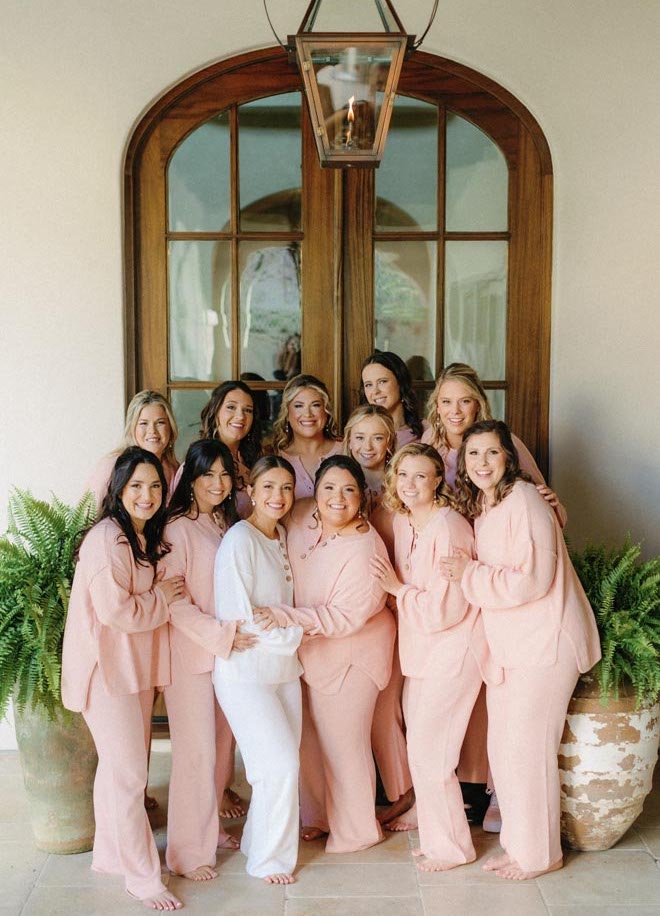 The bride and her bridesmaids smile in their peach getting ready outfits for the wedding. 