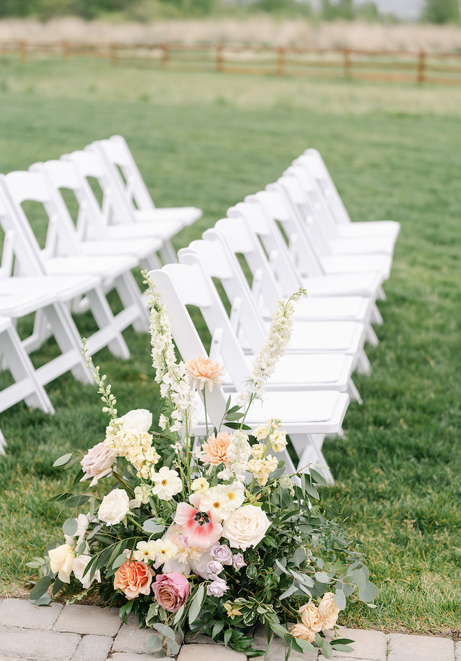 A floral arrangement with pink and ivory florals with greenery on the end of a line of white chairs. 