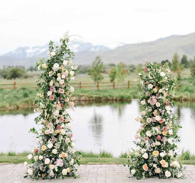 A floral installation of greenery, ivory, purple and pink florals with a pond and mountains behind it. 