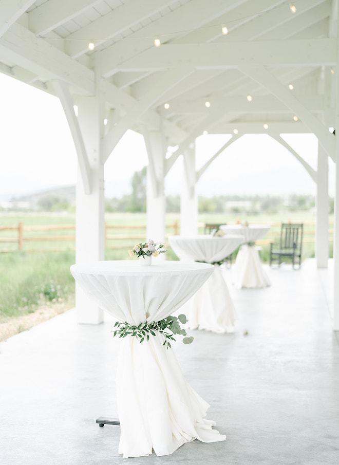 White cocktail tables wrapped with greenery lining a patio in the white-barn venue. 