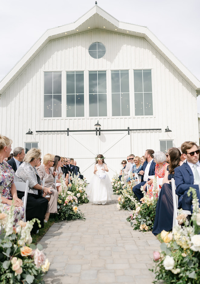 The flower girl walking down the aisle with the barn-style venue behind her. 