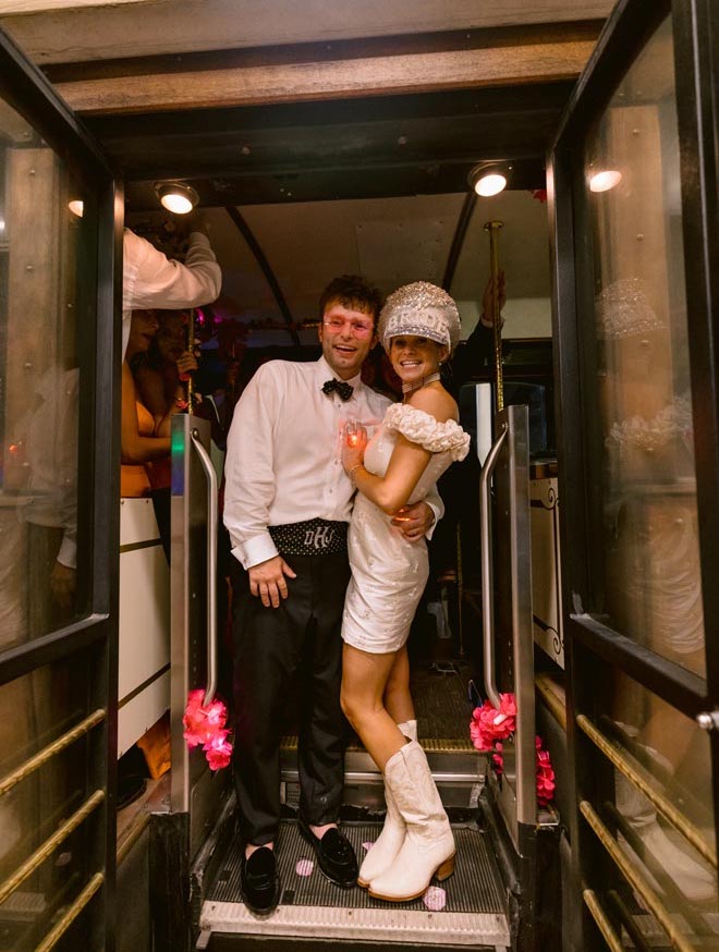 The bride and groom standing at the doorway in a trolley. The bride is wearing a mini dress, cowboy boots and a hat that says "Bride."