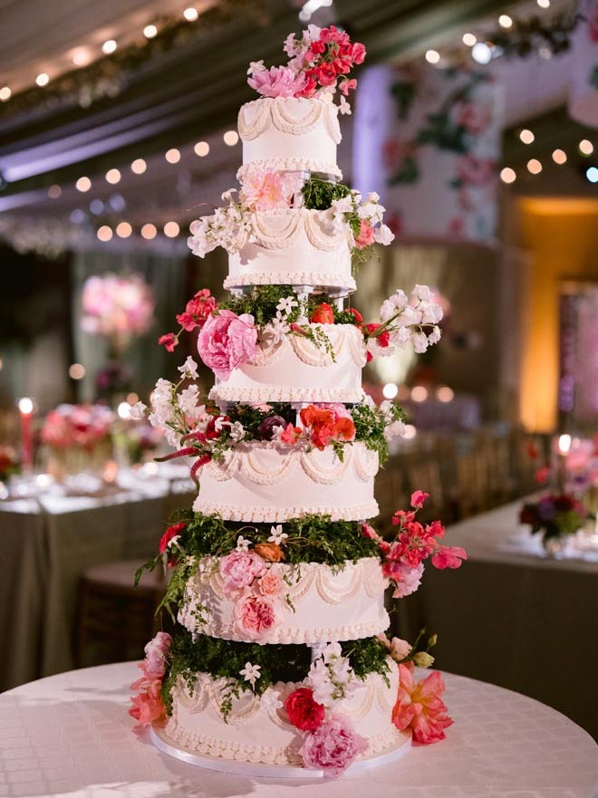 A six tier white cake with greenery in between each tier and garnished with pink and orange flowers. 