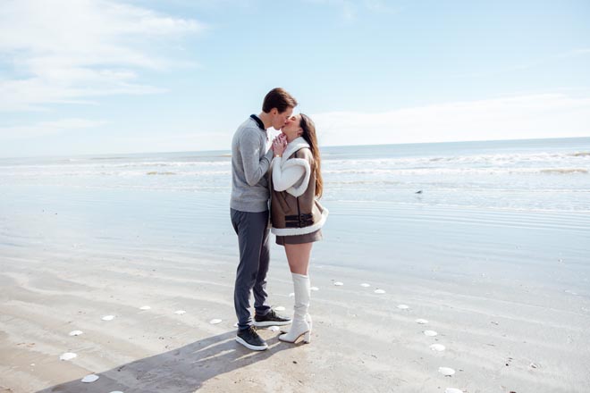 The couple kiss on the beach surrounded by seashells in the shape of a heart. 