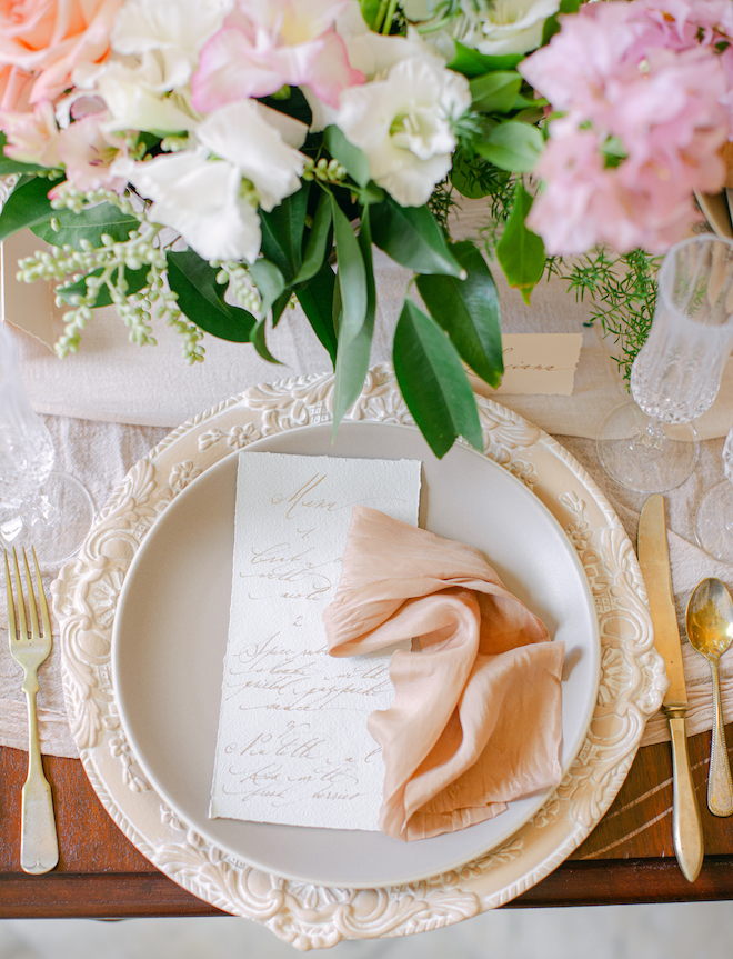 A table scape with a pink and white bouquet, beige plates and silver flatware with peach-colored linens. 
