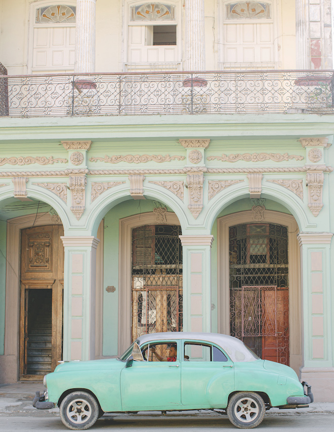 A vintage turquoise car parked against an old turquoise and white building in Havana. 