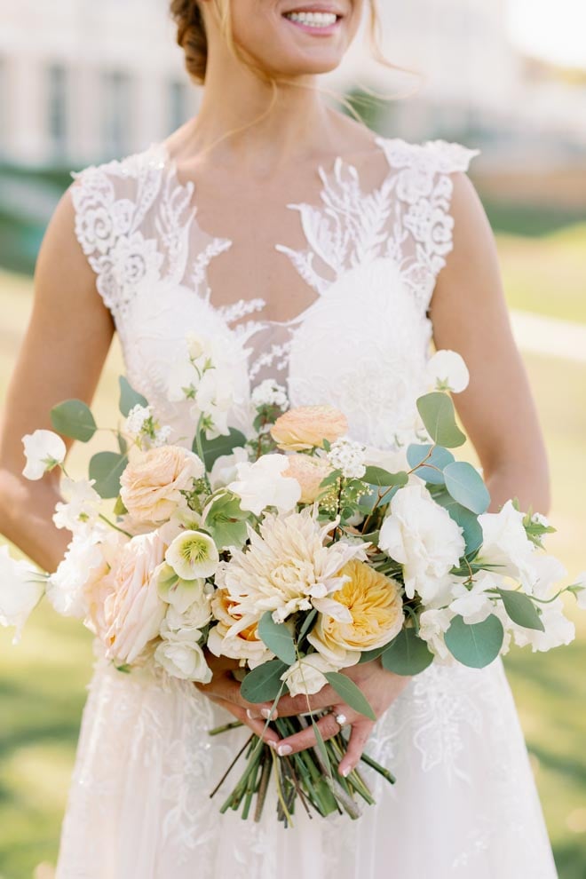 The bride holds her wedding bouquet of white, peach and green florals. 