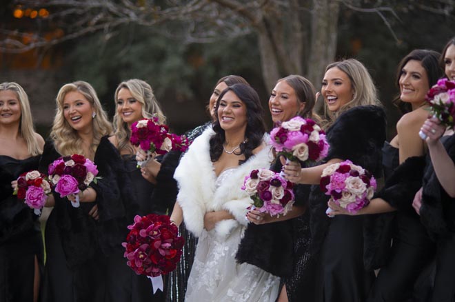 The bridesmaids wear black dresses and hold pink wedding bouquets while standing next to the bride. 