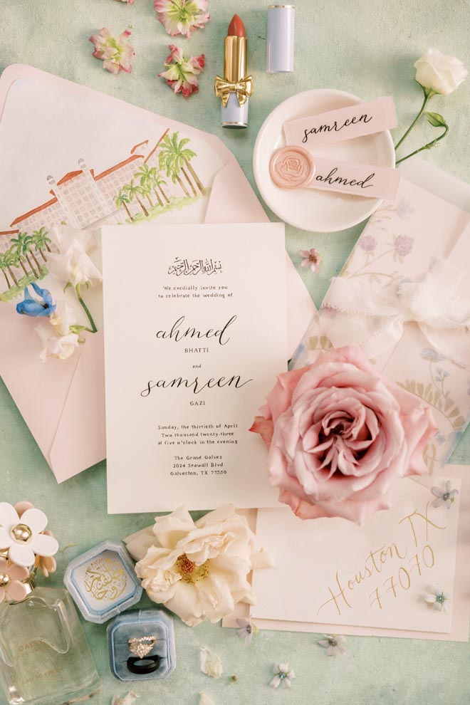 Pink pastel wedding stationery lay around the bride's jewelry and perfume. 