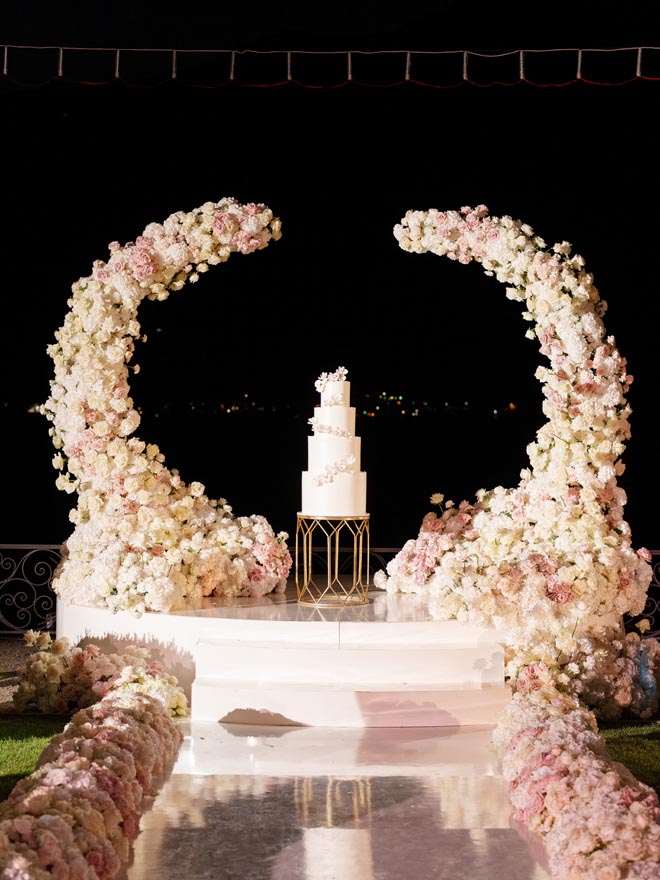 A four-tier white wedding cake is set under a blush floral arch. 