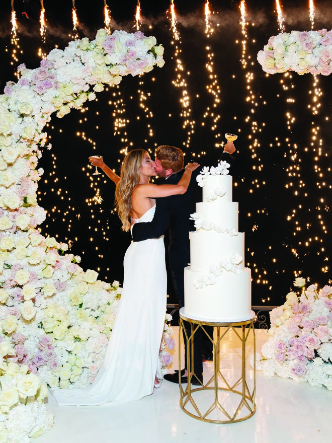 The bride and groom share a kiss next to their four-tier wedding cake and sparklers. 