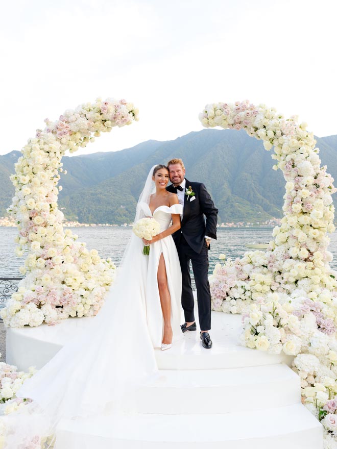 The couple pose under blush floral arch at their al fresco wedding on Lake Como. 