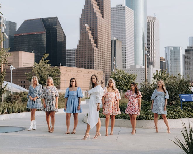 A bride in white with six girls in colorful dresses standing behind her against the Houston skyline. 