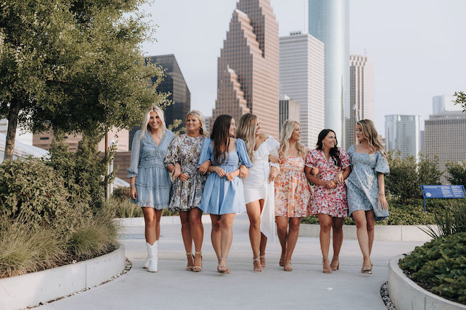 A bride in white with six bridesmaids in colorful sundresses standing hooking arms and walking against the Houston skyline. 
