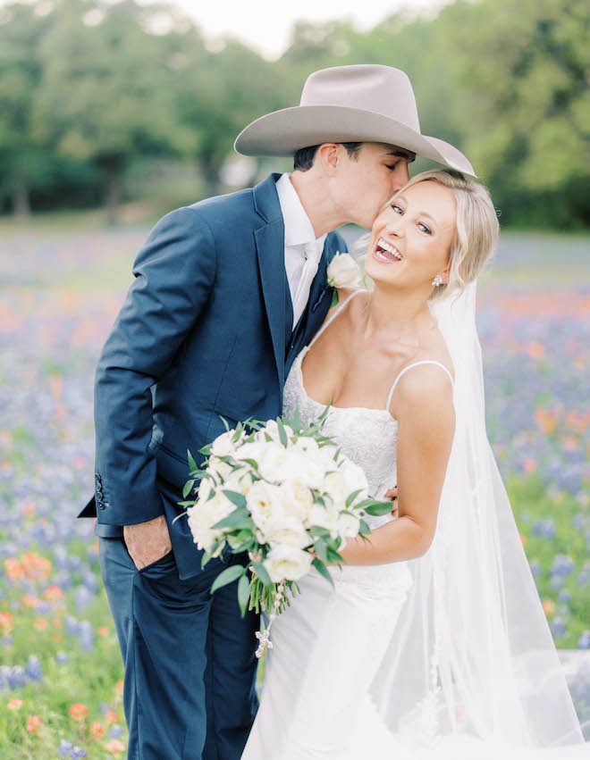 A groom wearing a cowboy hat kissing his bride's cheek in a field of bluebonnets. 