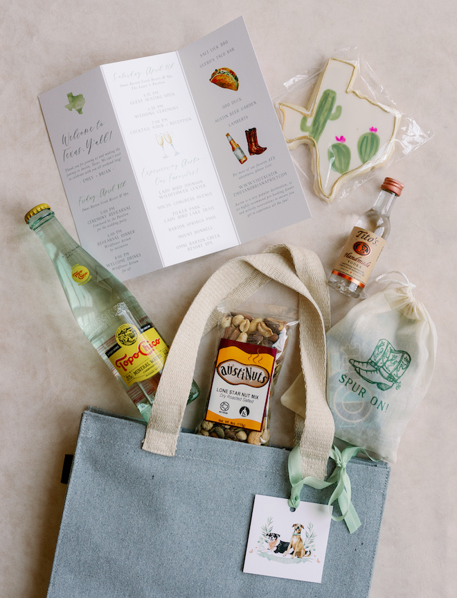 A blue canvas bag with a print of two dogs on the tag with a wedding itenerary, cactus painted cookie, topo chico bottle, Tito's Vodka shooter and Austi-Nuts. 