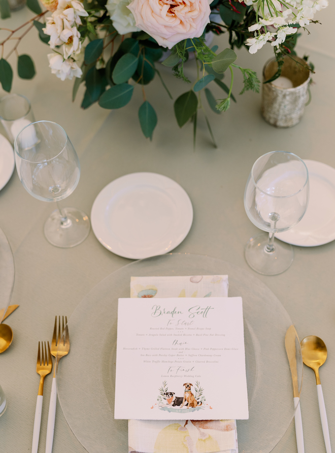 A tablescape with green linens, gold flatware an a custom menu with a print of a black and white dog and a brown dog at the bottom. 