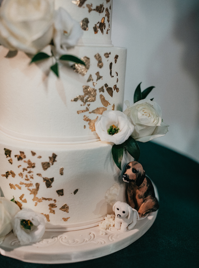 Three tier white cake with gold animal print and white florals with two cake-sculpted dogs made to look like they took a bite of cake. 