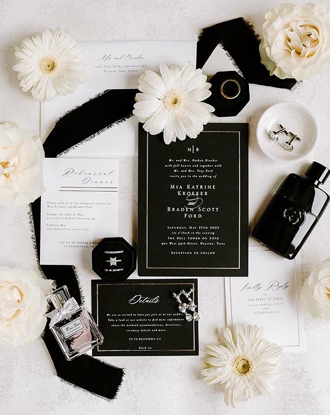 A black-and-white wedding invitation suite with white florals decorating the flatlay. 